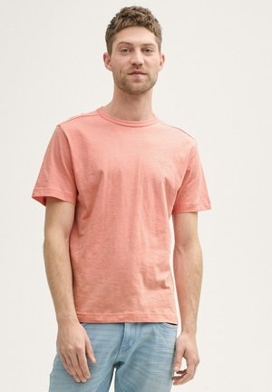 Jeune homme portant un t-shirt à manches courtes rose saumon et un jean bleu clair, se tenant devant un fond blanc uni.