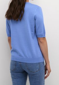 Donna con lunghi capelli castani che indossa un maglione azzurro a maniche corte e jeans di denim blu, vista da dietro su uno sfondo bianco.