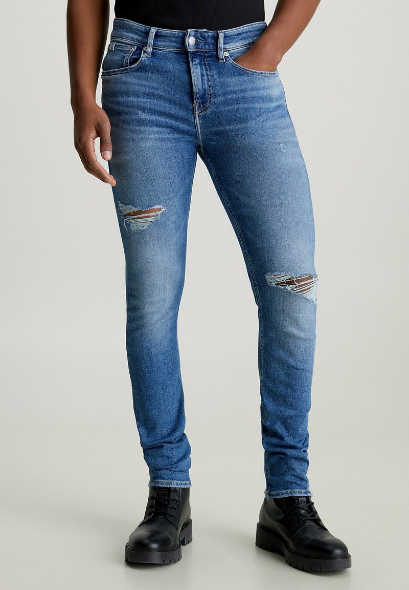 Person trägt eng anliegende, zerrissene blaue Jeans mit Abnutzungsspuren an den Oberschenkeln und schwarzen Schnürstiefeln, steht mit einer Hand in der Tasche.