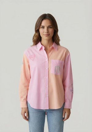 Donna che indossa una camicia con bottoni a righe verticali rosa e pesca con ricami e jeans azzurri, in piedi davanti a uno sfondo semplice.