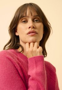 Maglione fucsia lavorato a maglia con scollo a V, polsini a costine e texture morbida, indossato su uno sfondo chiaro. Mano poggiata sul mento.