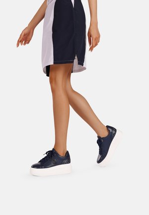 Sneakers blu navy con finitura lucida, suola a piattaforma bianca, design con lacci e dettagli borchiati sul tallone. Abbinati a una gonna sportiva.