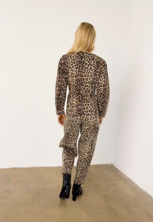 Femme aux cheveux blonds longs portant un haut à manches longues et un pantalon assorti imprimés léopard, bottines noires à talons, debout face à un mur blanc.