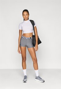 Λευκό crop top με κοντά μανίκια, γκρι σορτς με μαύρη ζώνη στη μέση, διάφανα αθλητικά παπούτσια και λευκές κάλτσες. Το μοντέλο κρατά μια μαύρη τσάντα. Απλό φόντο.