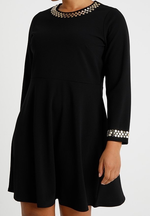 Schwarzes langärmeliges Kleid mit runder Halslinie und Manschetten, die mit metallischen runden Nieten verziert sind, getragen von einer Person mit entspanntem Arm an der Seite.