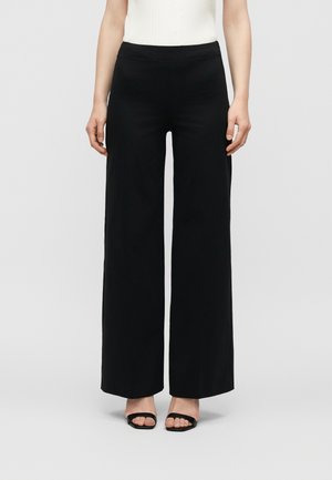 Persoană purtând pantaloni largi negri cu talie înaltă, asortați cu pantofi negri decupați la vârf și un top alb fără mâneci.