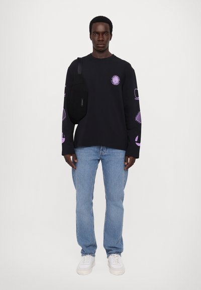 Schwarzes Langarmshirt mit lila Grafiken, kombiniert mit hellblauen Jeans und weißen Sneakers. Die Umhängetasche wird an der linken Seite getragen.