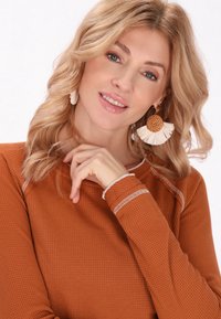 Hosszú ujjú, textúrált felső égett narancssárga színben, fehér varrási díszítéssel. A modellen nagy, kerek, szőtt fülbevalók láthatók, bojt részlettel.