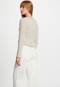 Mujer con suéter de punto beige con puños abotonados y pantalones blancos de cintura alta, posando mirando por encima del hombro contra un fondo blanco.