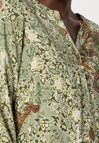 Grüne Bluse mit floralem Muster und Knopfleiste, die aufwendige Paisleymuster in Orange- und Gelbtönen zeigt. Weicher, strukturierter Stoff.