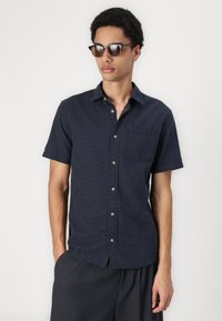 Chemise à manches courtes bleu marine à boutons avec un motif texturé, poche poitrine et col classique, portée avec un pantalon ample de couleur sombre.