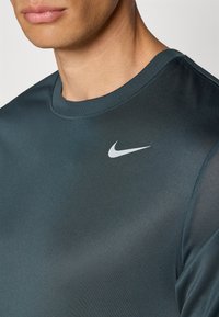Nike atletisk skjorte i mørk teal, med en glat tekstur, korte ærmer og et kontrastfarvet hvidt swoosh-logo på venstre bryst.