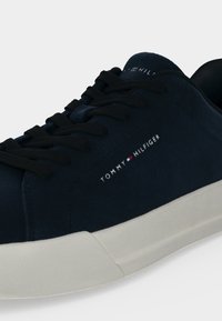 Σκούρο μπλε αθλητικό παπούτσι Tommy Hilfiger με μαύρα κορδόνια και λευκή σόλα, με το όνομα της μάρκας και το λογότυπο στο πλάι και στη γλώσσα.