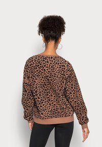 Mujer con cabello rizado recogido en un moño alto, lleva pendientes de aro, un suéter de estampado leopardo en marrón y negro, y pantalones negros ajustados, vista desde atrás.