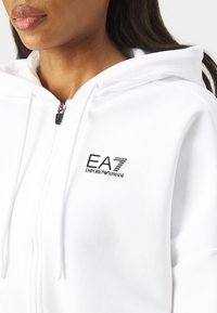 Bela zip-up kapuca narejena iz mehkega blaga, z black EA7 Emporio Armani logotipom na levi prsni strani in vrvico za zategovanje na kapuci.