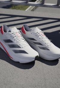 Zapatillas deportivas blancas con rayas negras y detalles rojos, colocadas sobre una superficie de hormigón con patrones de sombra proyectados por una barandilla encima.