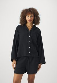 Schwarzes Baumwoll-Loungewear-Set mit einem locker sitzenden Hemd mit Knopfleiste und einer Tasche sowie passenden Shorts. Strukturierter Stoff und weiche Oberflächen.