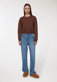 Giovane donna che indossa un maglione marrone, jeans blu larghi e scarpe slip-on marroni, in piedi su uno sfondo bianco uniforme.
