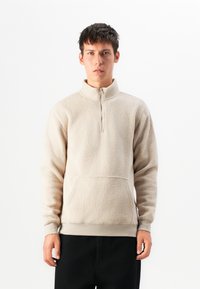 Beige fleece pullover med en halvlukket krave, tekstureret overflade, forlommer og ribbede manchetter, parret med sorte bukser.