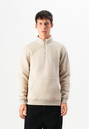 Beige fleece pullover met een halve ritssluiting, textuur oppervlak, een voorzak en geribbelde manchetten, gecombineerd met zwarte broeken.