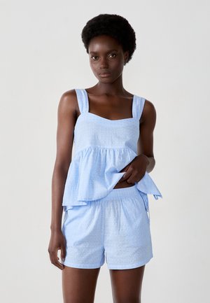 Top peplum in cotone azzurro chiaro con spalline larghe, motivo a texture e pantaloni corti abbinati. Presenta una vita elastica e lacci laterali per regolazione.
