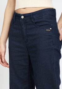 Dunkelblaue Jeans aus Denim mit mittlerer Bundhöhe, ausgestattet mit einem silbernen Knopfverschluss und einem kleinen schwarzen Markenetikett an der Vordertasche.