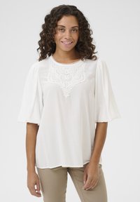 Blusa bianca con maniche corte a sbuffo e un dettaglio in pizzo sullo scollo. Tessuto morbido con una vestibilità rilassata e un orlo curvo.