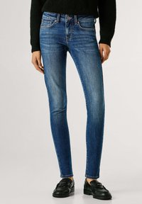 Õhukesed sinised teksad, mis on valmistatud denimist, keskmise vöökohaga, õrna tuhmumisega ja klassikalise viie taskuga ning kombineeritud mustade loaferitega.