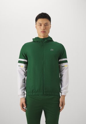 Veste de sport verte avec capuche, dotée de manches blanches, d'une fermeture éclair à l'avant, et du logo Lacoste noir sur la poitrine et le bas. Textură de tissu lisse.
