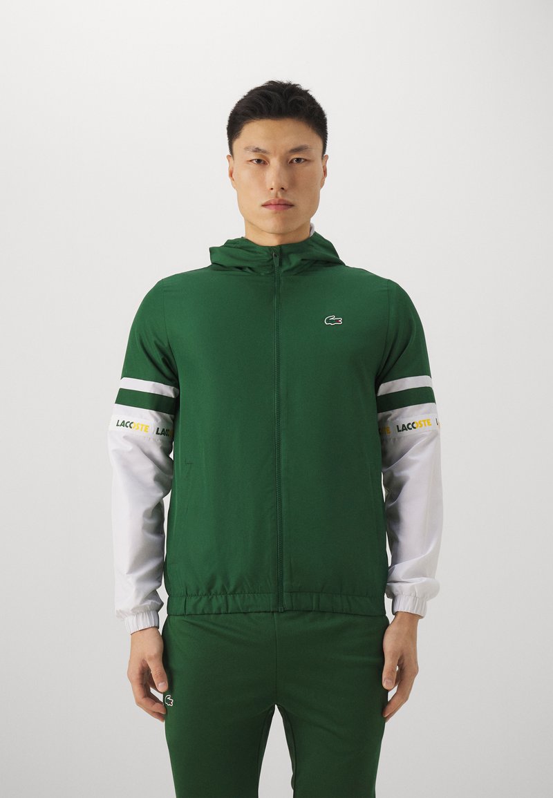 Grüne Sportjacke mit Kapuze, weiße Ärmel, durchgehender Reißverschluss und schwarzes Lacoste-Logo auf der Brust und den Hosen. Glatte Stoffoberfläche.