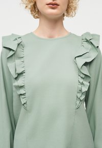 Γυναίκα που φοράει ανοιχτό πράσινο μακρυμάνικο μπλουζάκι με διακοσμητικές βολάν στους ώμους και στο μπροστινό μέρος.