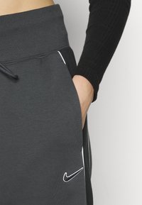 Gros plan sur une personne portant un pantalon de jogging Nike gris foncé avec une main dans la poche et un haut à manches longues noires côtelé.