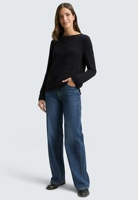 Pull en maille noire avec un col bateau et des manches évasées, associé à un jean en denim bleu taille haute et des chaussures noires.