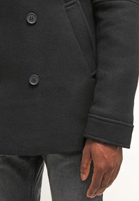 Svart ullkappa med fyra knappar, slagkrage och en vågrät söm på ärmen, tillsammans med mörka jeans.