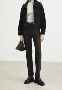 Chaqueta negra corta con bolsillos de parche sobre una blusa gris, combinada con pantalones negros de tiro alto y botas negras de tobillo de estilo robusto, sosteniendo un bolso negro.