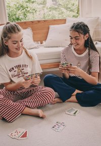 To piger sidder med benene krydsede på et lyst tæppe og holder farverige spillekort. Den ene har røde pyjamas med striber på, den anden blå. En træbænk står i baggrunden.