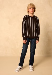 Brun randig tröja med vita vertikala linjer, kombinerad med mörkblå jeans och grå sneakers. Modellen står mot en neutral bakgrund.