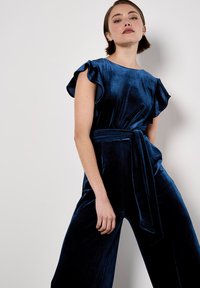 Mono de terciopelo azul marino con mangas cortas con volantes, cintura ceñida con cinturón y diseño de pierna ancha. Textura suave con una apariencia elegante y costuras mínimas.
