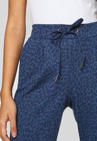 Femme portant un pantalon bleu à cordon avec un imprimé léopard bleu foncé et un haut blanc, la main reposant à côté du pantalon.