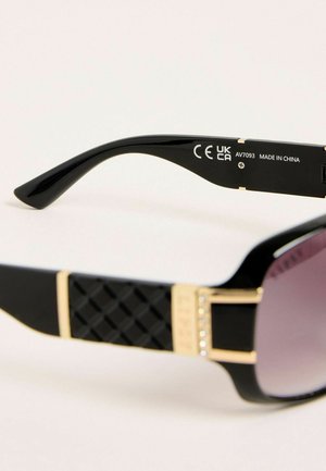 Lunettes de soleil surdimensionnées avec des montures en plastique noir, un côté texturé noir et un accent doré ornées de petites pierres claires. Les verres ont un dégradé sombre.