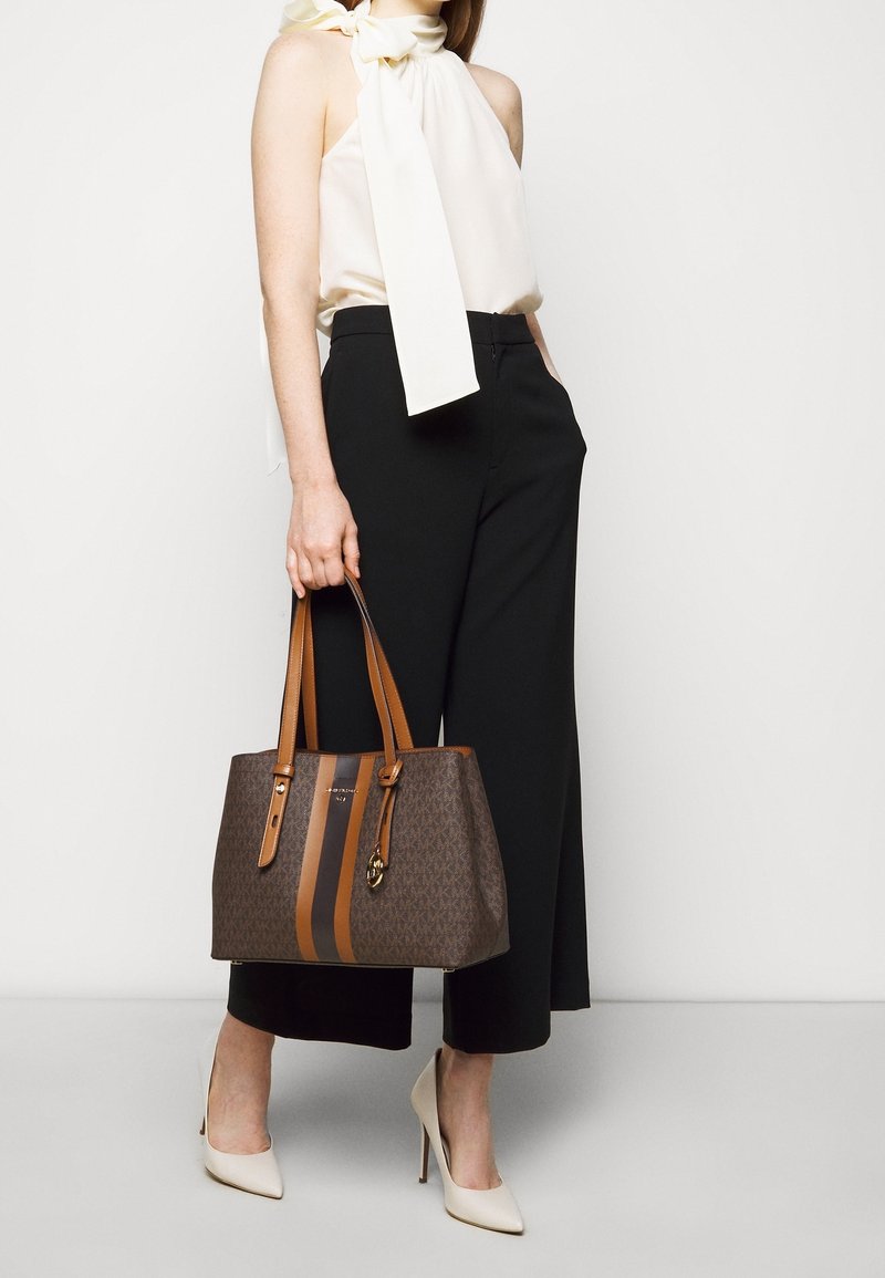 Femme en blouse blanche sans manches avec un nœud au col, pantalon noir à jambes larges, talons blancs, tenant un sac cabas marron avec des rayures beige.