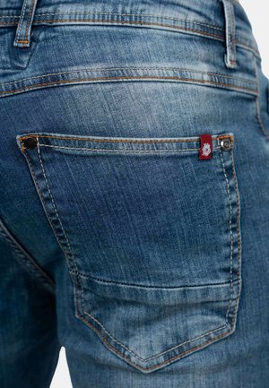 Denim jeans in een medium blauwe was, met oranje stiksels, een achterzak met een rood label en een gladde textuur.