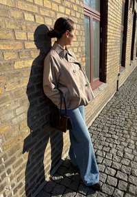 Beżowa dwurzędowa marynarka, szerokie niebieskie dżinsy, brązowa torebka, na tle teksturowanej ściany z cegły, z cieniami rzucanymi na kamienną nawierzchnię.