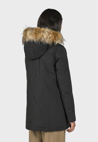 Svart parka-jacka med en lurvig huva i tan och brunt, med dragkedjestängning, långa ärmar och knälång design. Slät, matt yta.