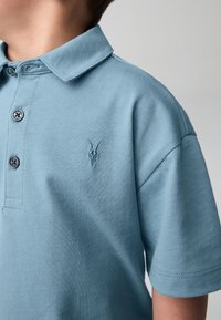 Ljusblå bomullspikétröja med uppvikt krage, tre knappslå och en liten broderad logotyp på bröstet. Mjuk textur.