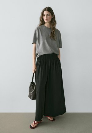 Donna con lunghi capelli che indossa una maglia grigia a maniche corte, pantaloni neri a gamba larga, tiene in mano una borsa nera e sta scalza su un pavimento grigio davanti a una parete chiara e liscia.