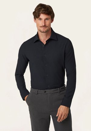 Man die een aansluitend zwart, lange mouwen, knoopssluiting overhemd draagt, ingestopt in grijze nette broek, met één hand in zijn zak staand.