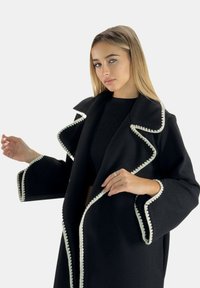 Black wool coat with contrasting white scalloped trim, deep collar, open front, oversized sleeves, and ribbed black shirt underneath.