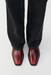 Persona che indossa jeans scuri e scarpe quadrate prugna lucide, in piedi su un pavimento bianco, vista dall'alto.