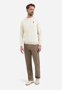 Lichtcrème trui met ronde hals en klein zwart logo, gecombineerd met bruine broek en witte sneakers. Zachte textuur en casual pasvorm.